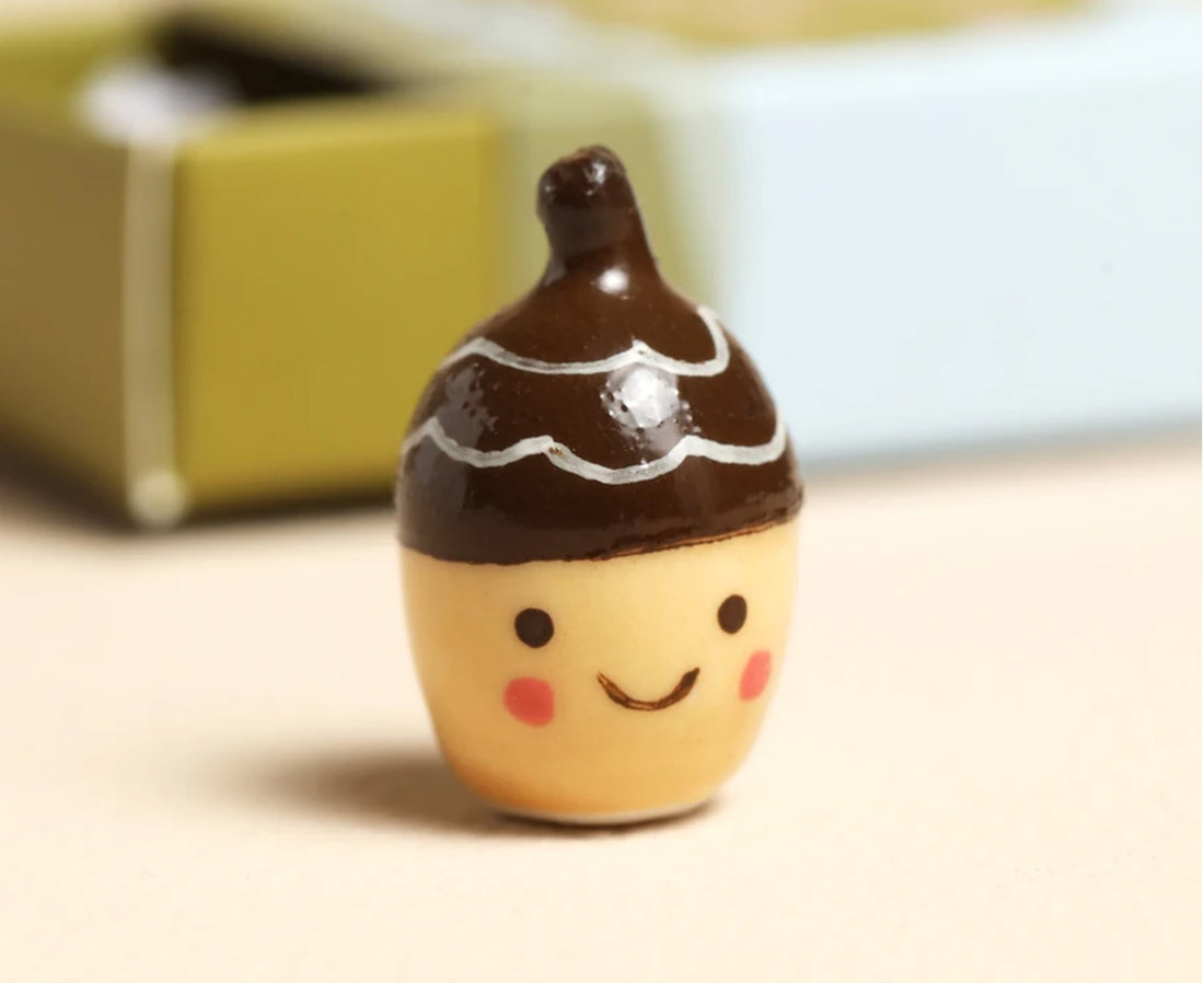A cute, small acorn-shaped figurine with a smiling face, featuring a dark brown top with white icing-like stripes and a warm beige bottom, set against a softly blurred background.