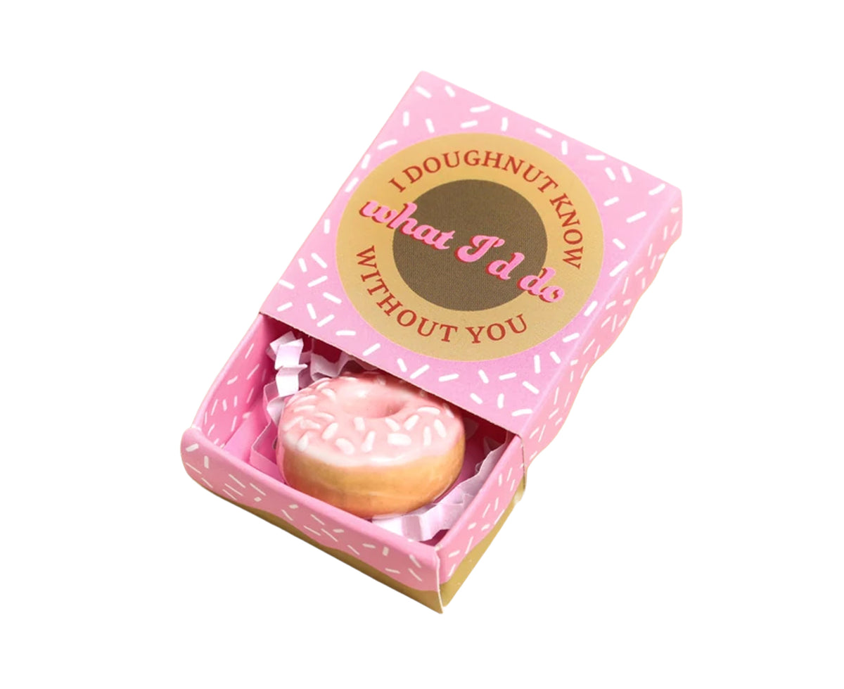 A pink matchbox-style gift box with a donut-shaped item inside, shown on a plain white background. The lid has a gold oval label reading &