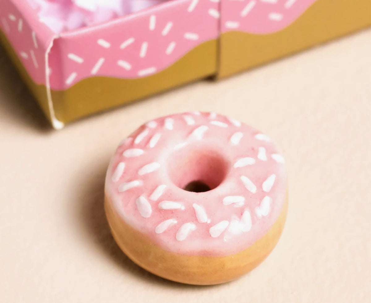 A miniature pink frosted donut with white sprinkles, shown in sharp focus in the foreground with a blurred pink and gold box in the background.