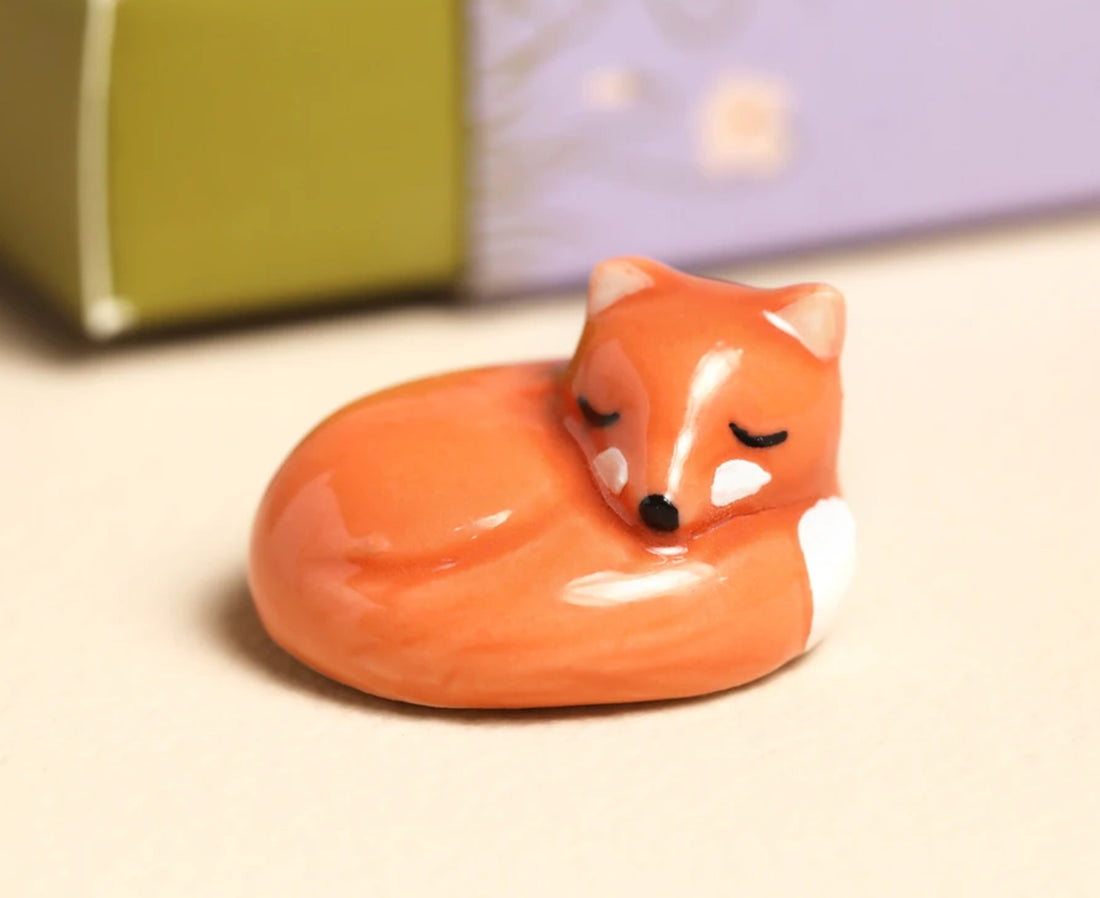 A small glossy ceramic fox figurine curled up sleeping, with orange, peach, white, and black details, set against a softly blurred background.