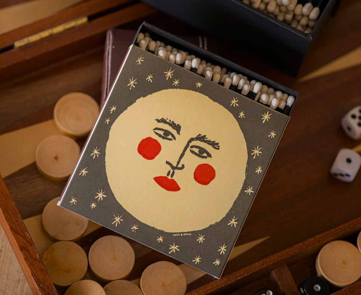 A decorative matchbox featuring a stylized moon with face surrounded by small gold stars on a dark charcoal background. The matches are shown with a game board and dice.