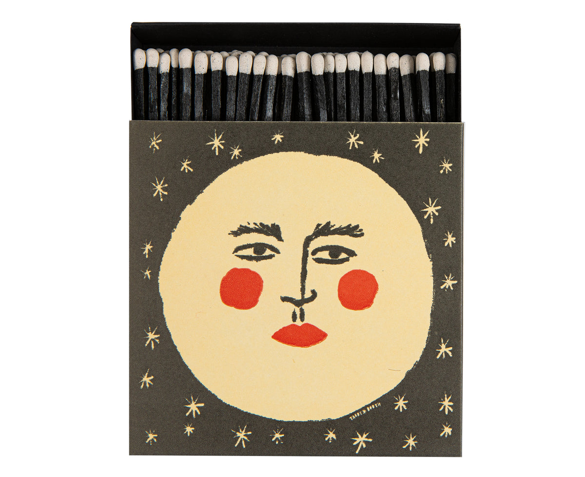 A decorative matchbox with black matches inside. The front cover features a stylized moon with face surrounded by small gold stars on a dark charcoal background.