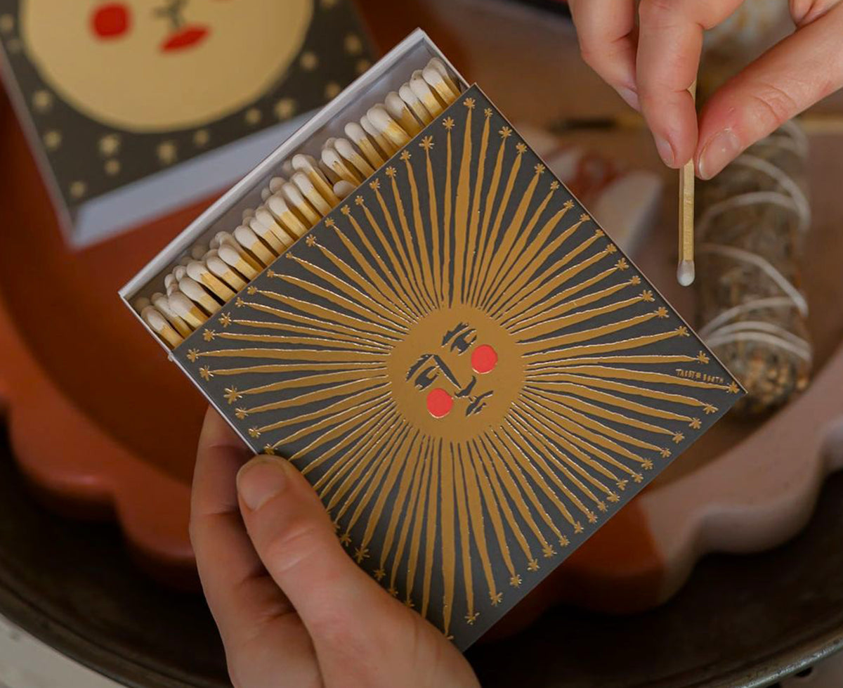 A matchbox with a black cover featuring a metallic gold sun design and red cheeks open and being held.