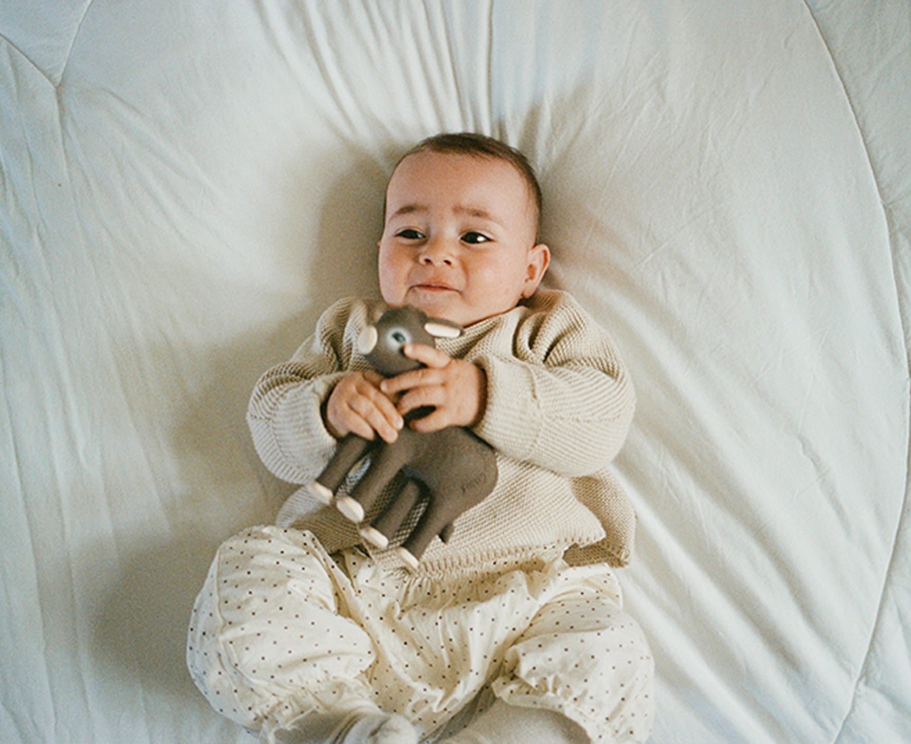 A baby lying on a white quilted mattress holding Hector the Donkey Chewable Toy by Oli & Carol