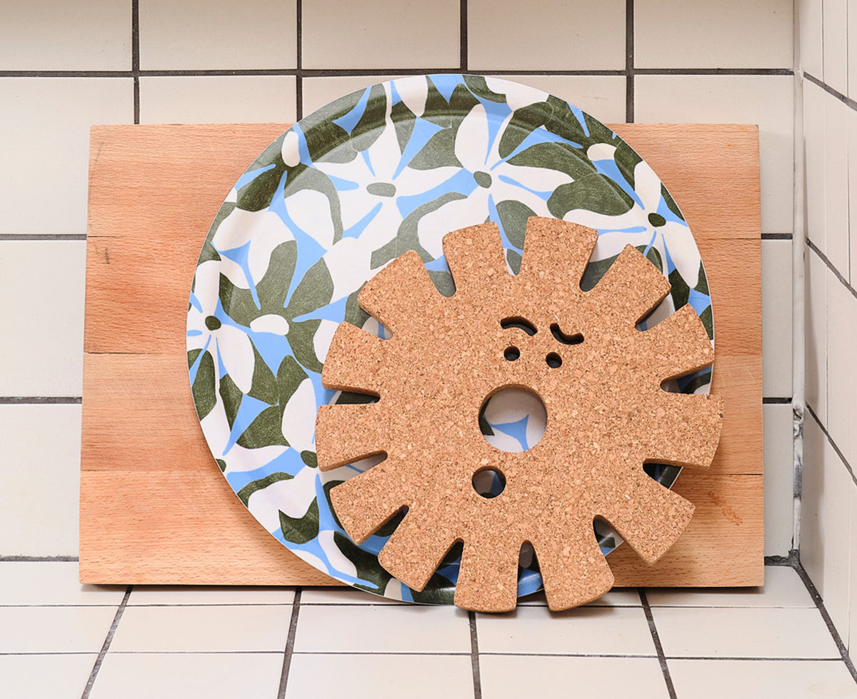 Sun Face Medium Cork Trivet by Wrap on a kitchen counter with a tray in the background.