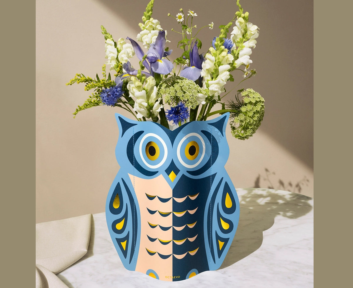 An paper vase featuring an owl design in blue, peach and gold by Octaevo. the vase is holding flowers and sits on a marble table.
