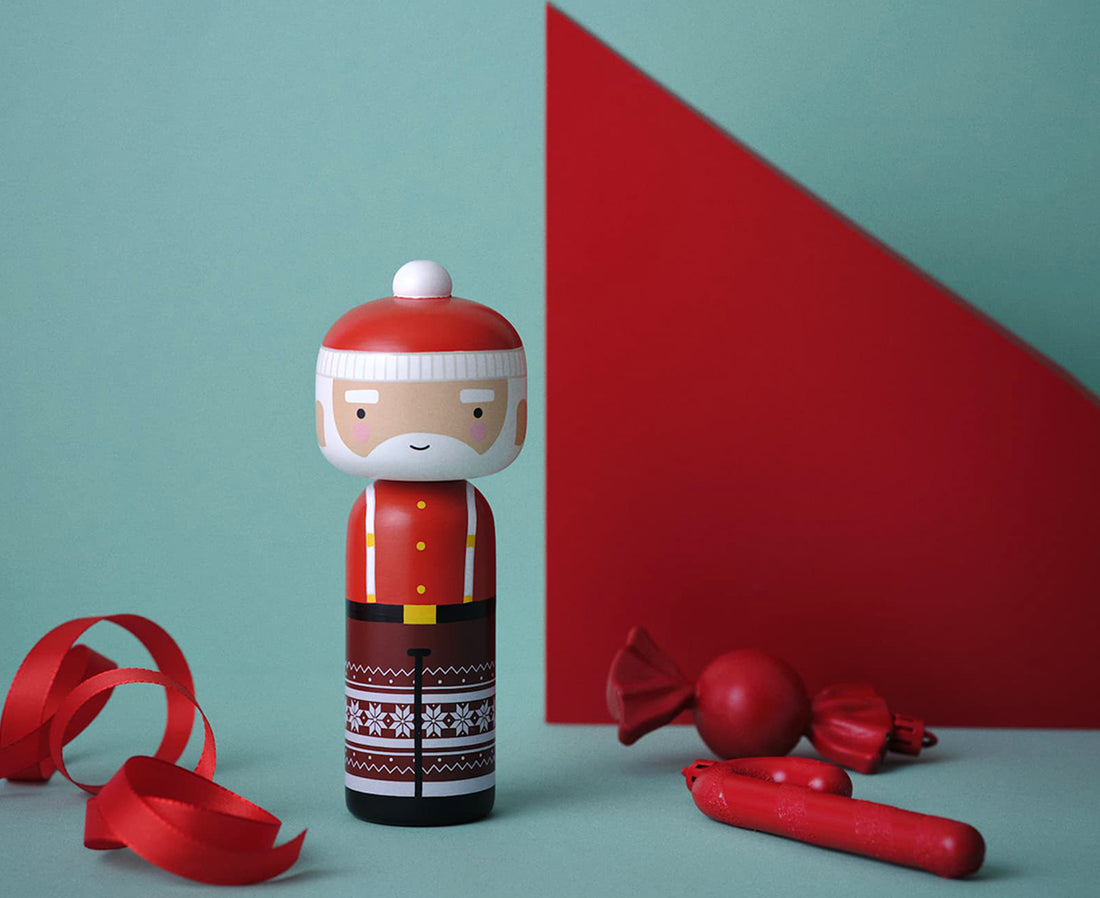 A Santa Claus-themed wooden kokeshi doll with a red hat, white beard, red jacket with yellow buttons, and a brown patterned skirt with white snowflakes, on a red and teal background.