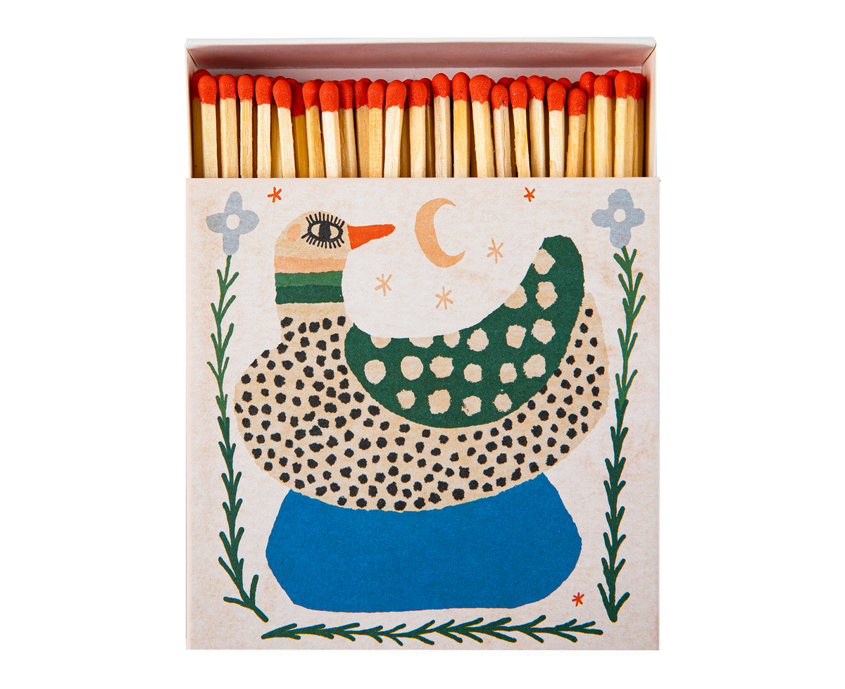 A decorative matchbox filled with wooden matches topped with red tips. The matchbox lid features a stylized spotted duck illustration in green, blue, black, and cream.