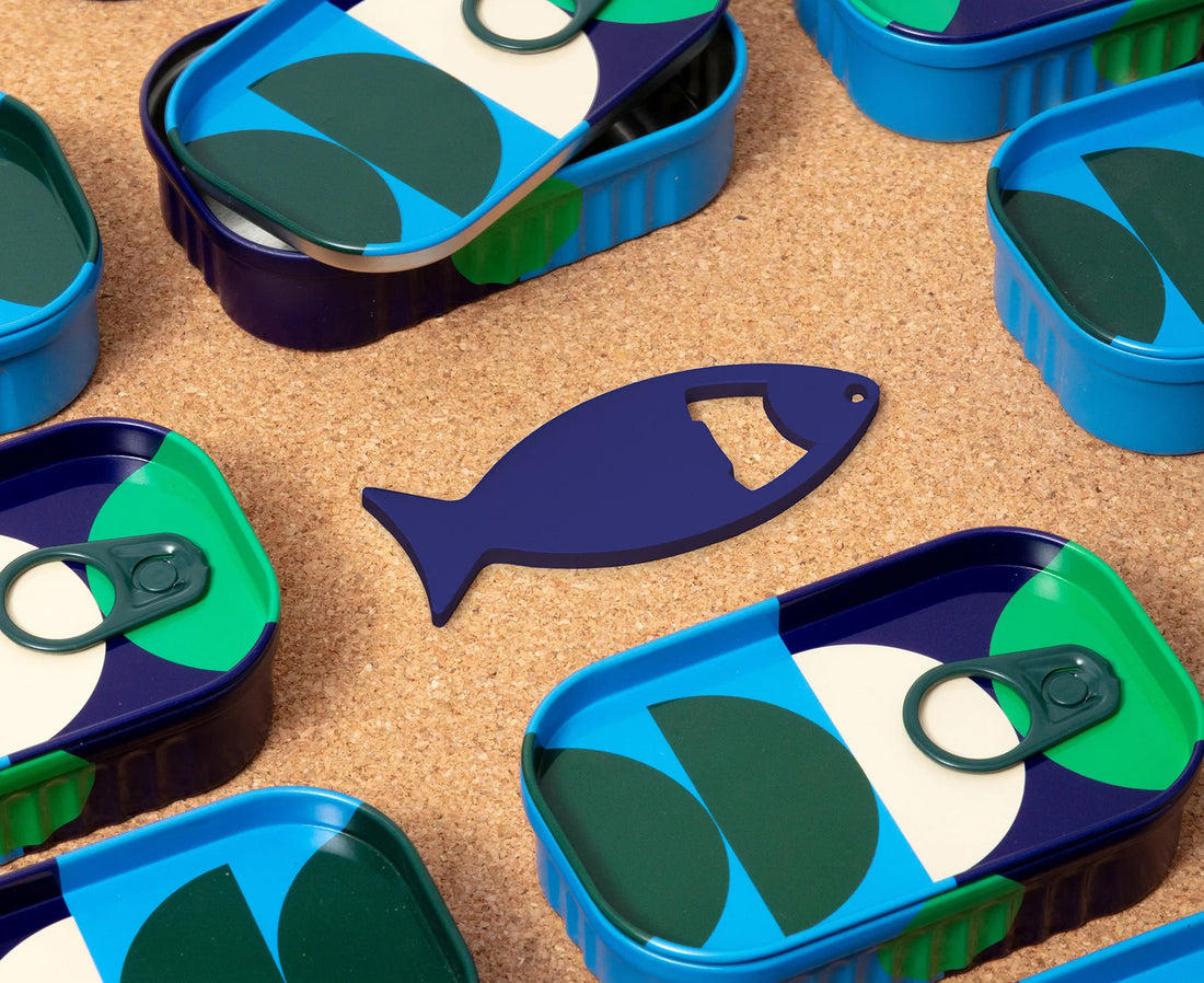 A blue fish-shaped bottle opener shown next to pull-tab cans with a geometric pattern of green, blue, beige, and dark purple on a cork background.