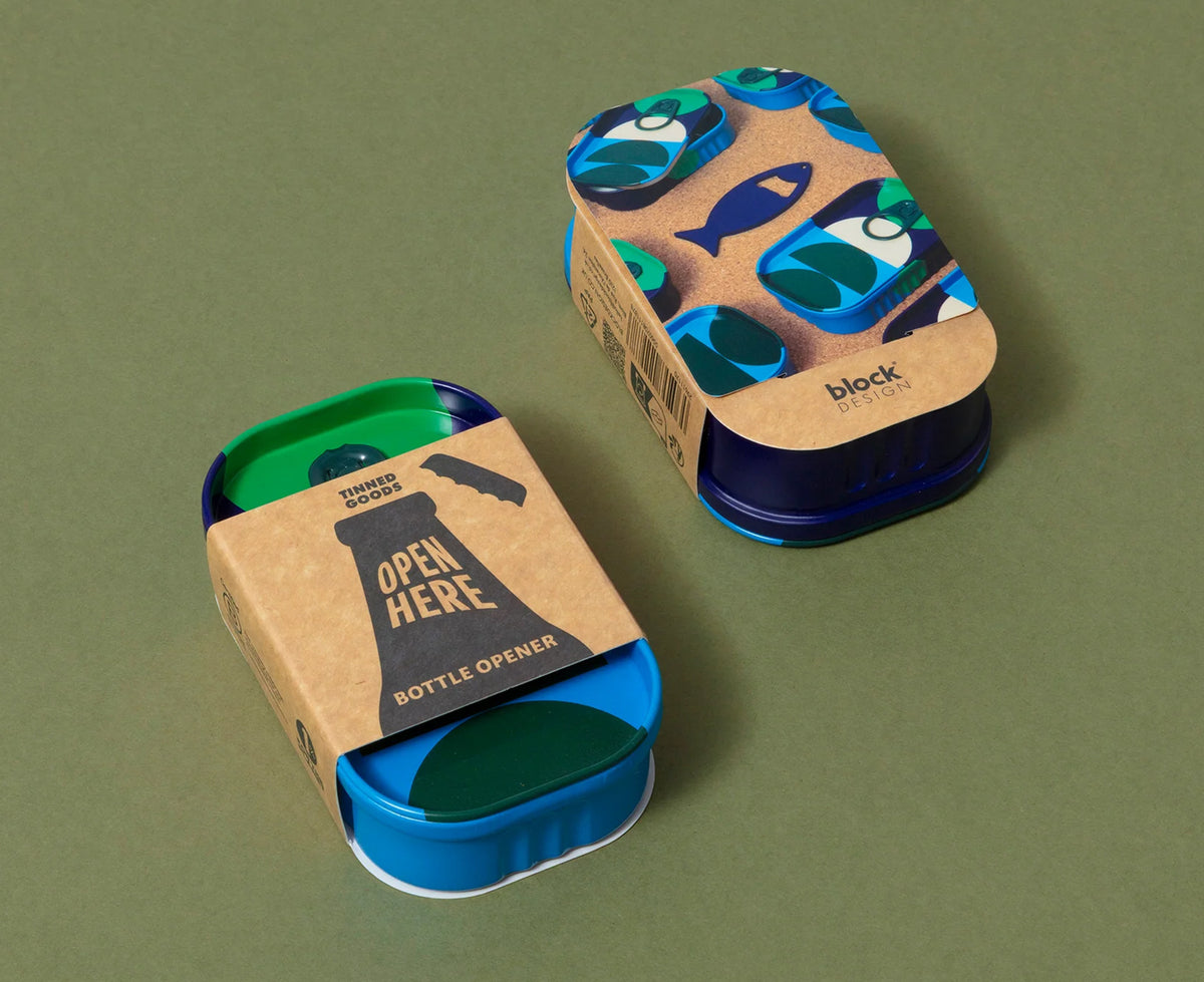 Two metal bottle openers on a muted olive green background. One open opener shows a blue base, green interior, and kraft paper sleeve with a black fish silhouette and the words &