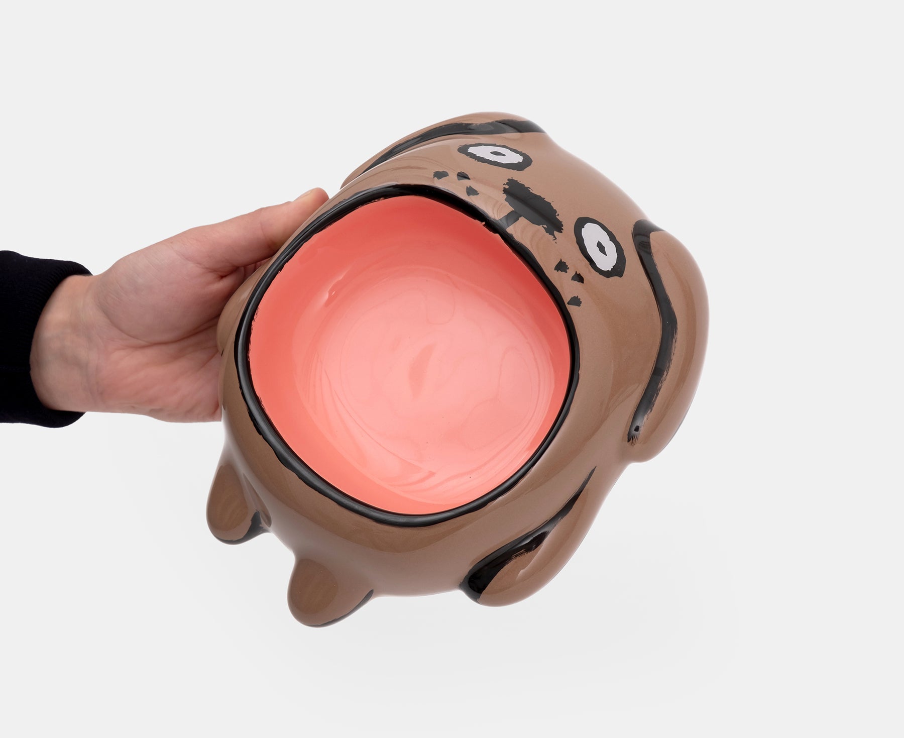 A ceramic Dog Bowl by Home Studyo with a brown body, black hand-drawn face, and a large glossy peach opening and hand.