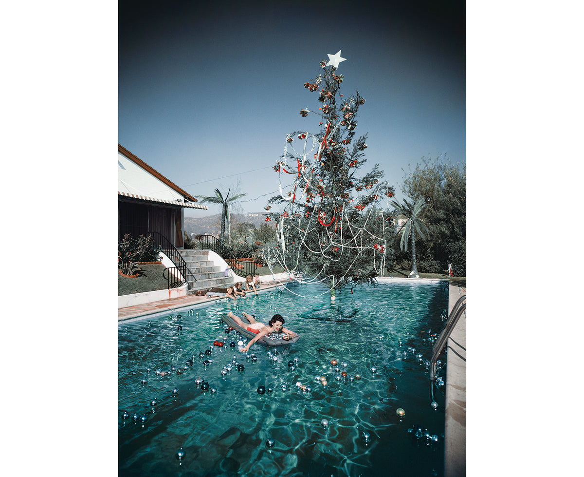 The artwork of the Christmas Swim puzzle features a person floating on an inflatable ring in a swimming pool beside a large, decorated Christmas tree with a star on top, with a house and palm trees in the background.