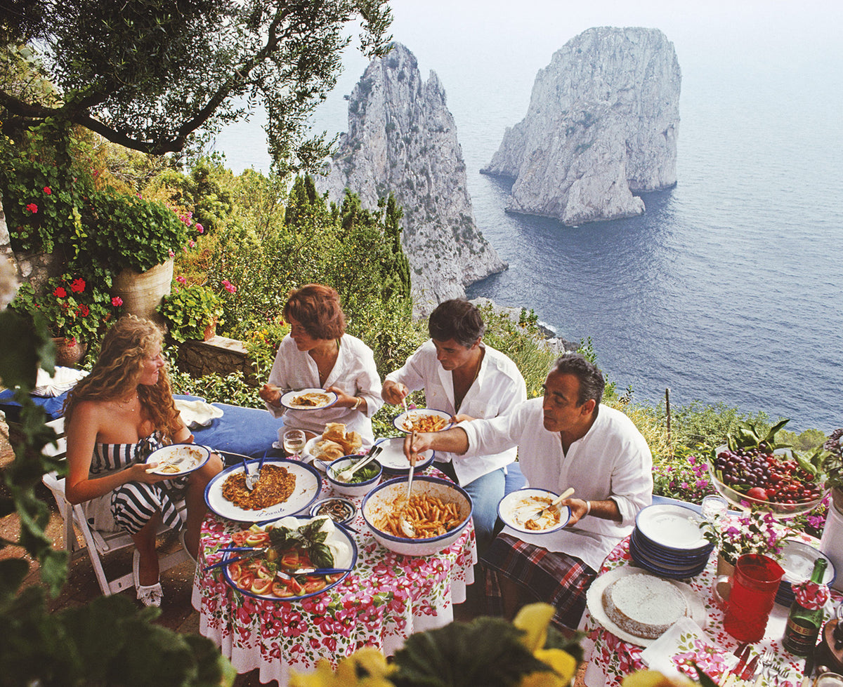 The artwork of the Dining Al Fresco on Capri shows people dining outdoors on a table set with plates of food, flowers, and a red floral tablecloth, overlooking a dramatic sea cliff landscape with large rock formations. 