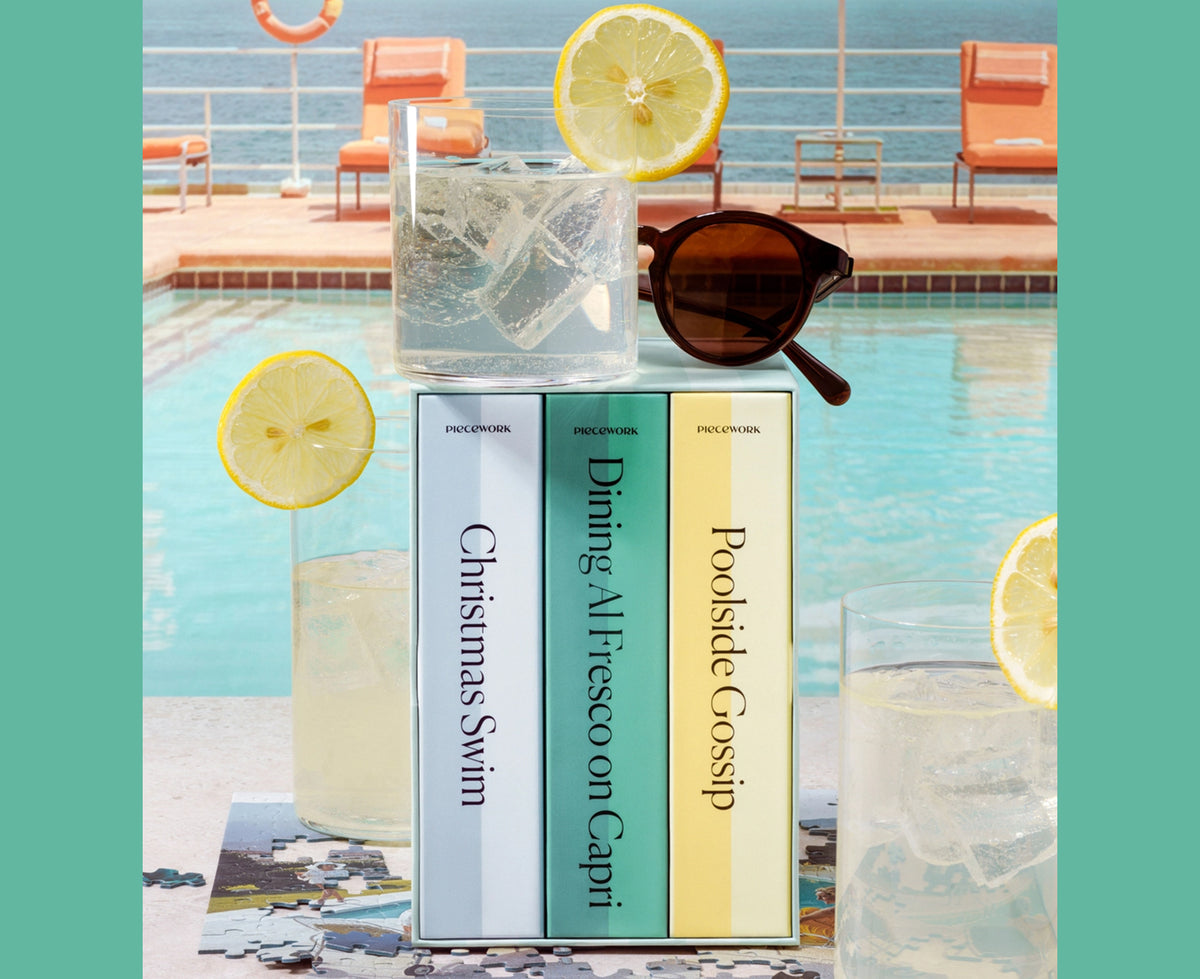 Three Piecework puzzle boxes with pastel blue, mint green, and pale yellow covers stand on a poolside surface, styled with glasses of iced tea, lemon slices, sunglasses, and a puzzle in a sunny outdoor setting.