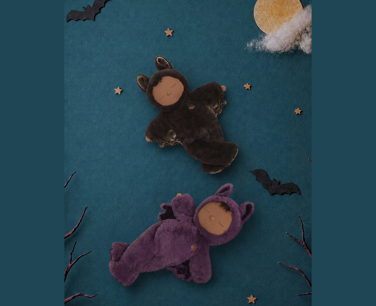 Two plush bat-shaped cozy dinkum toys, one dark brown and one purple, displayed on a dark teal background with decorative paper stars, bats, and a moon.