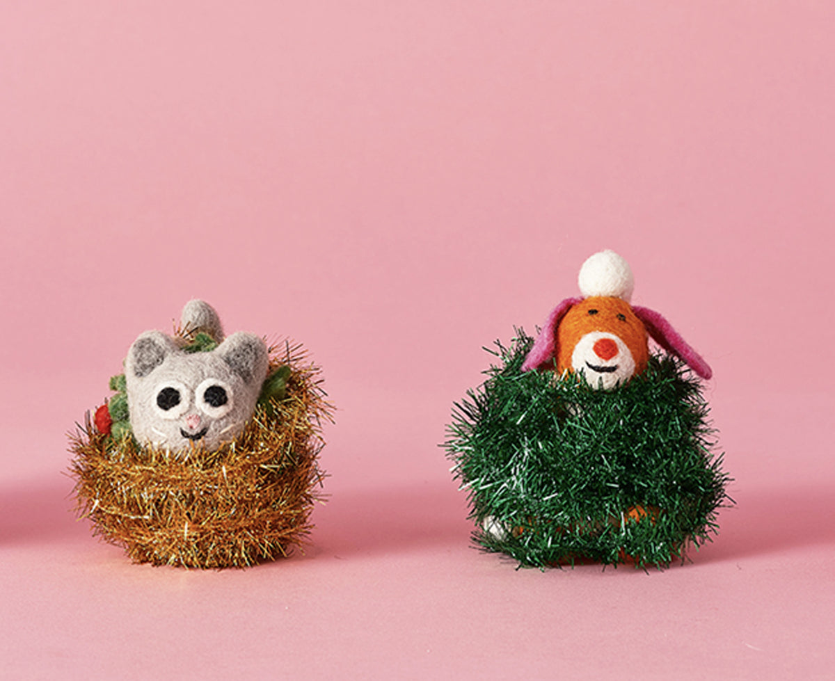 Two small handmade felt animal ornaments sitting in tinsel nests on a solid pink background. 