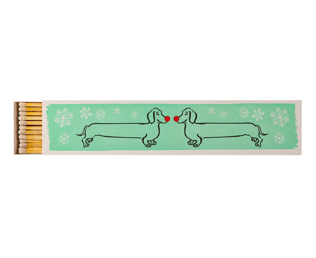 A long rectangular matchbox with a mint green printed panel showing two dachshund figures facing each other with small red noses, surrounded by white snowflakes. 