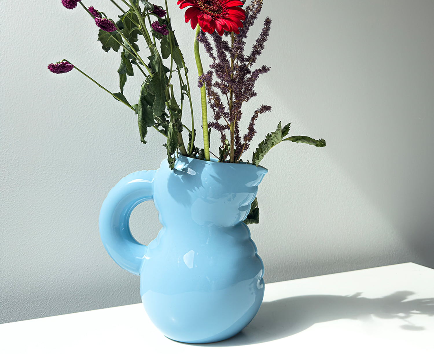 A light blue Blow Up ceramic pitcher by Home Studyo with a rounded, sculptural design, scalloped edges, and a curved handle holding flowers.