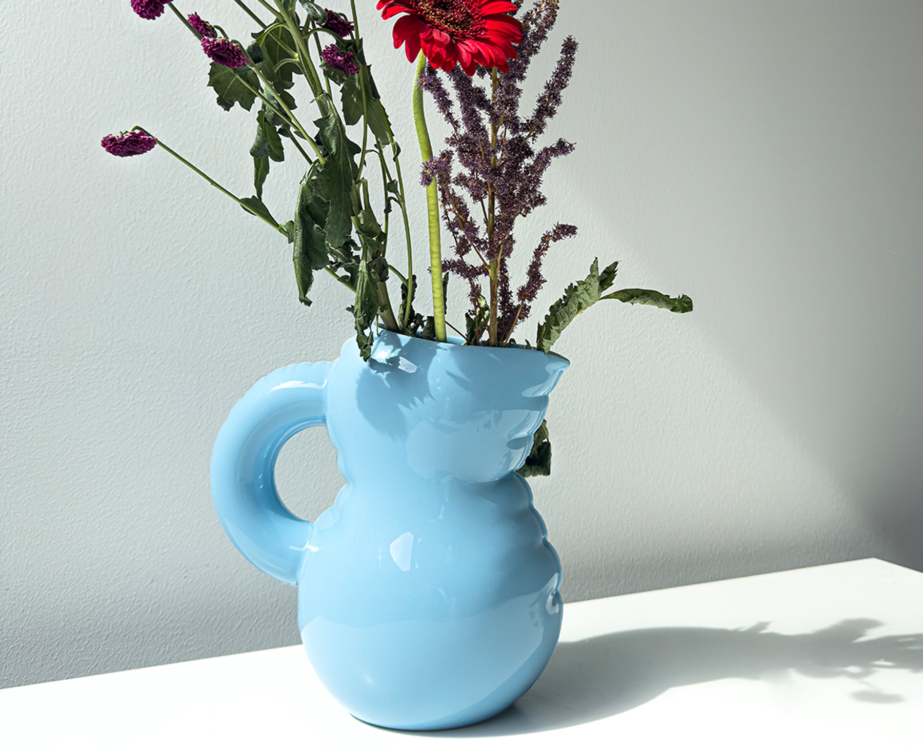 A light blue Blow Up ceramic pitcher by Home Studyo with a rounded, sculptural design, scalloped edges, and a curved handle holding flowers.