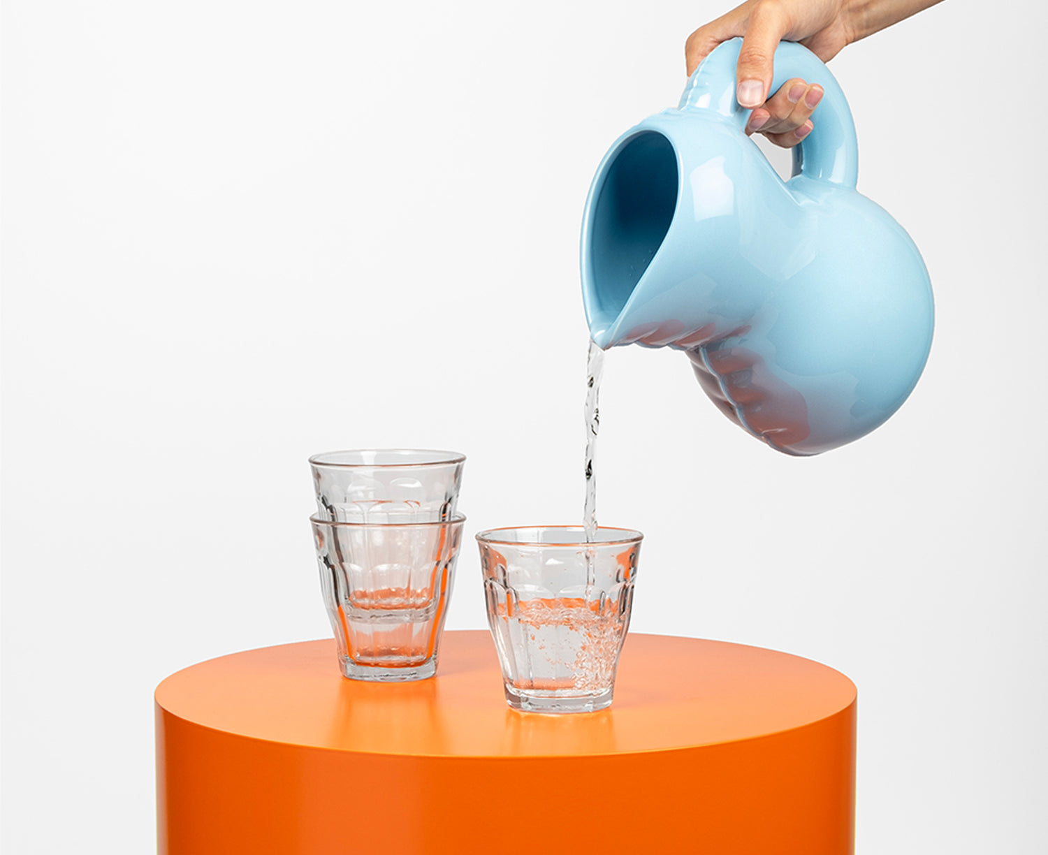 A light blue Blow Up ceramic pitcher by Home Studyo with a rounded, sculptural design, scalloped edges, and a curved handle pouring water.