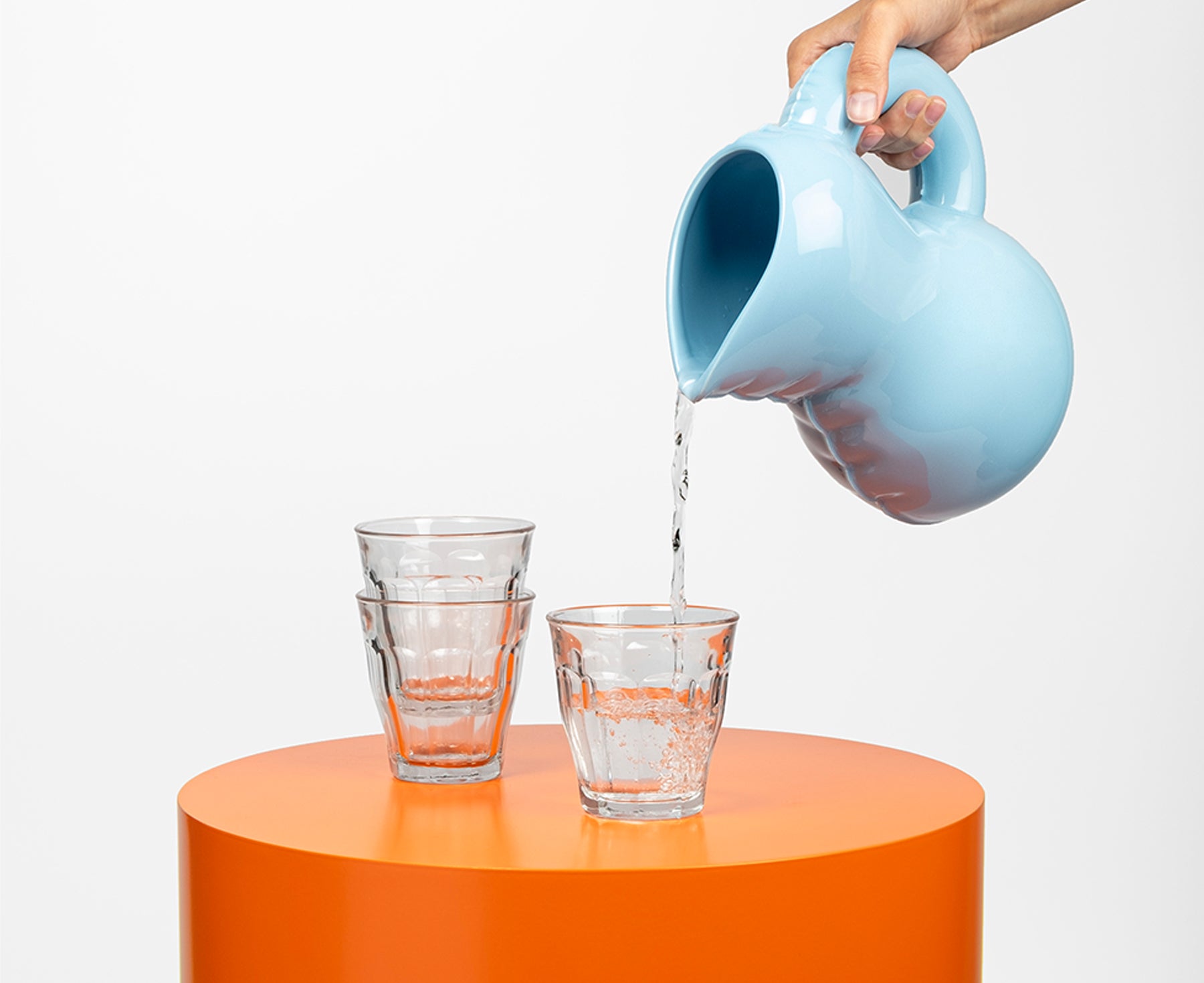 A light blue Blow Up ceramic pitcher by Home Studyo with a rounded, sculptural design, scalloped edges, and a curved handle pouring water.