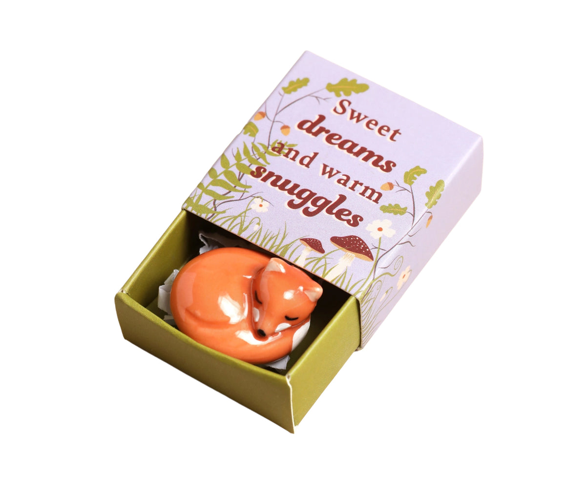 A small orange fox figurine nestled inside an open decorative gift box with a lavender lid printed with the phrase &