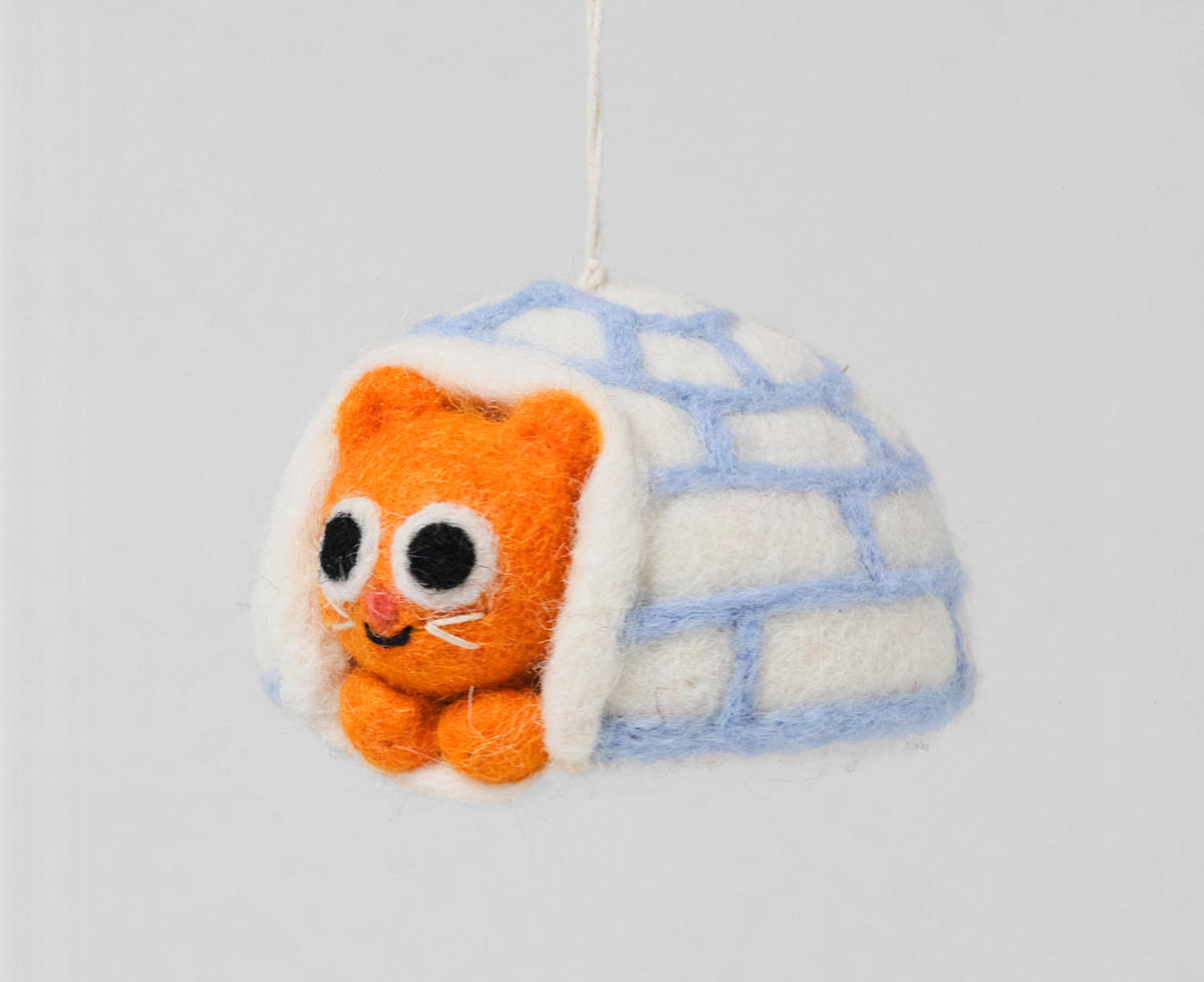 A felt hanging ornament depicting an orange kitten peeking out from a white igloo with light blue brick lines, suspended by a white string against a plain white background.