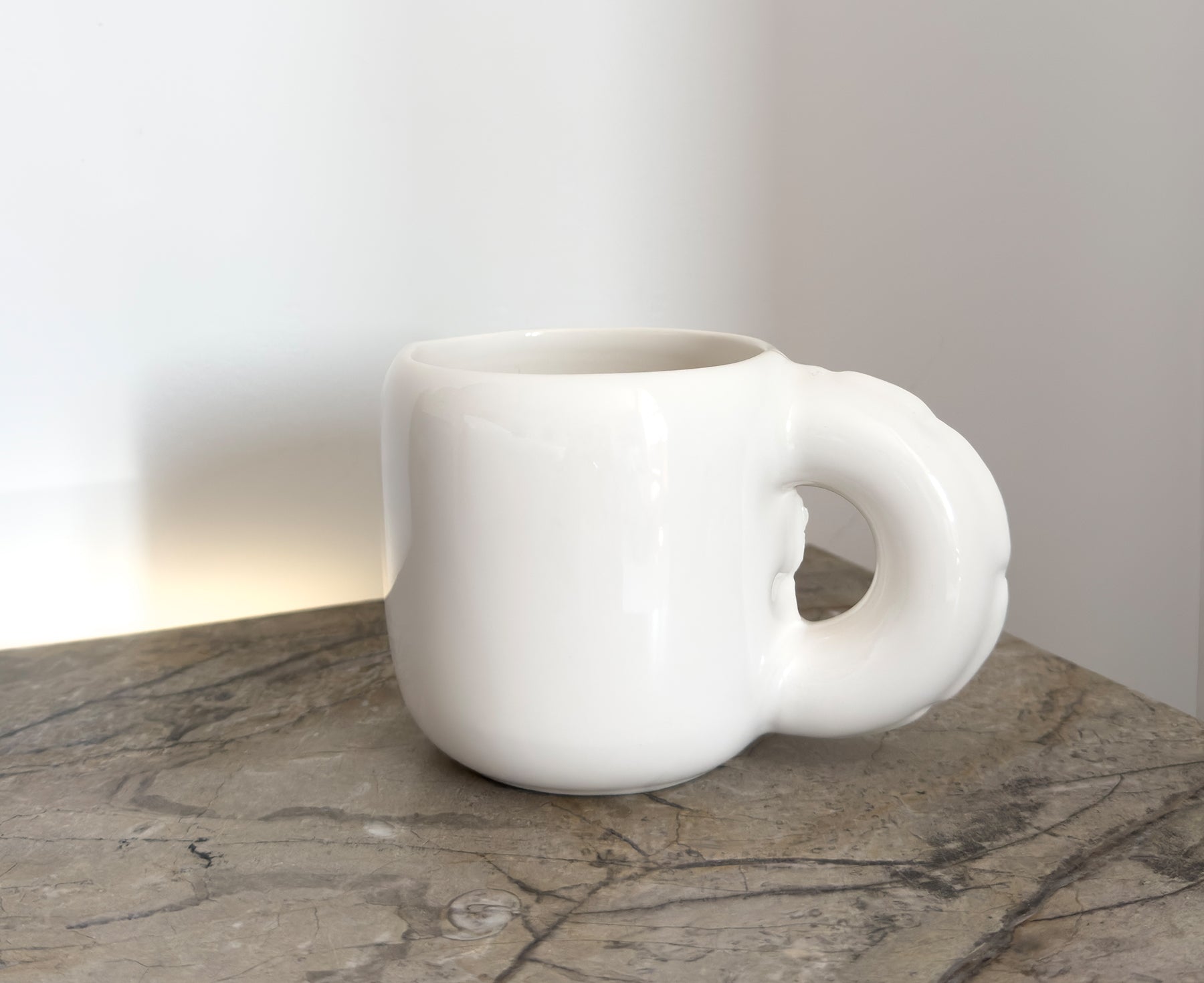 A glossy white ceramic Gaston Blow Up Mug by Home Studyo with a sculpted, organic handle and side details on a table.