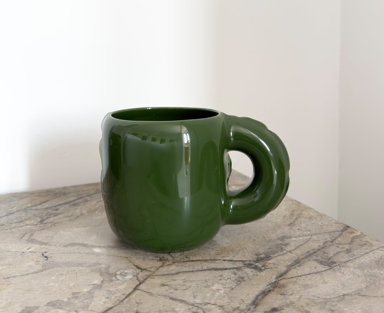 A glossy dark green ceramic Gaston Blow Up Mug by Home Studyo with a sculpted, organic handle and side details on a table.