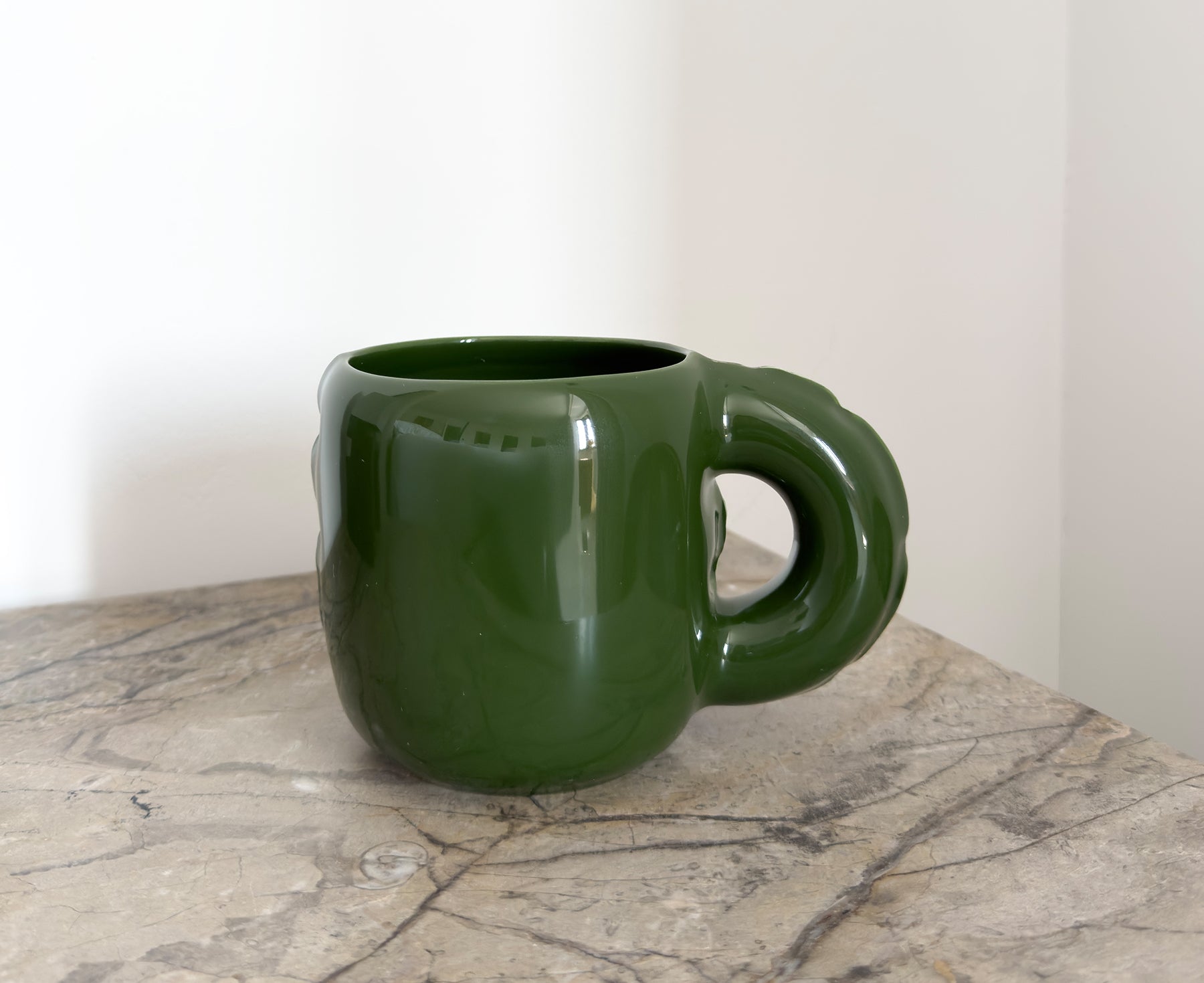 A glossy dark green ceramic Gaston Blow Up Mug by Home Studyo with a sculpted, organic handle and side details on a table.