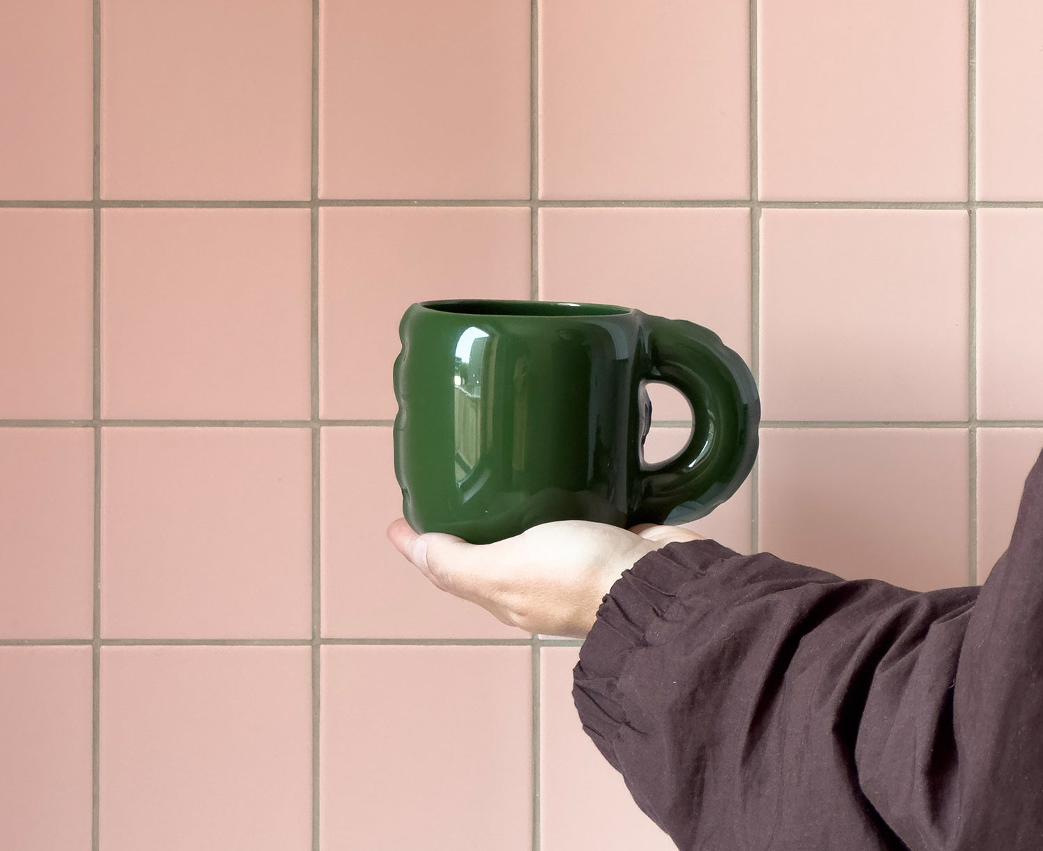 A glossy dark green ceramic Gaston Blow Up Mug by Home Studyo with a sculpted, organic handle and side details in a hand.