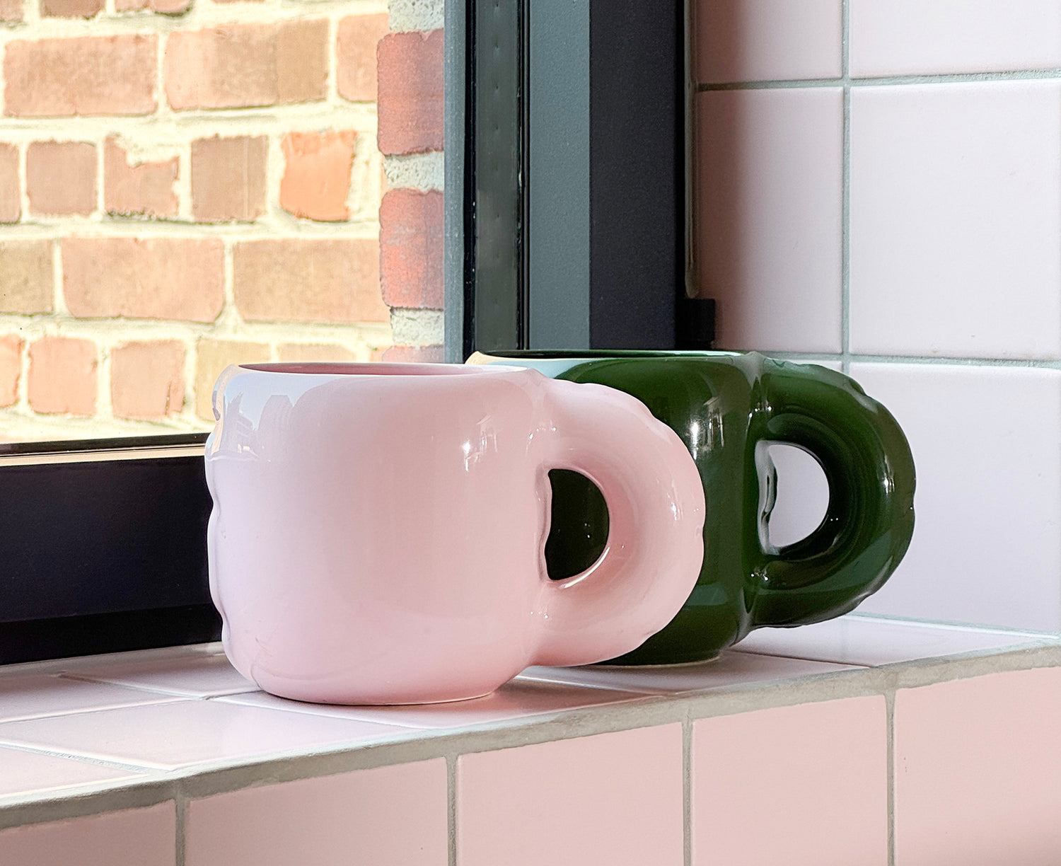 Glossy pink and green ceramic Gaston Blow Up Mug by Home Studyo with a sculpted, organic handle sitting on a window ledge.