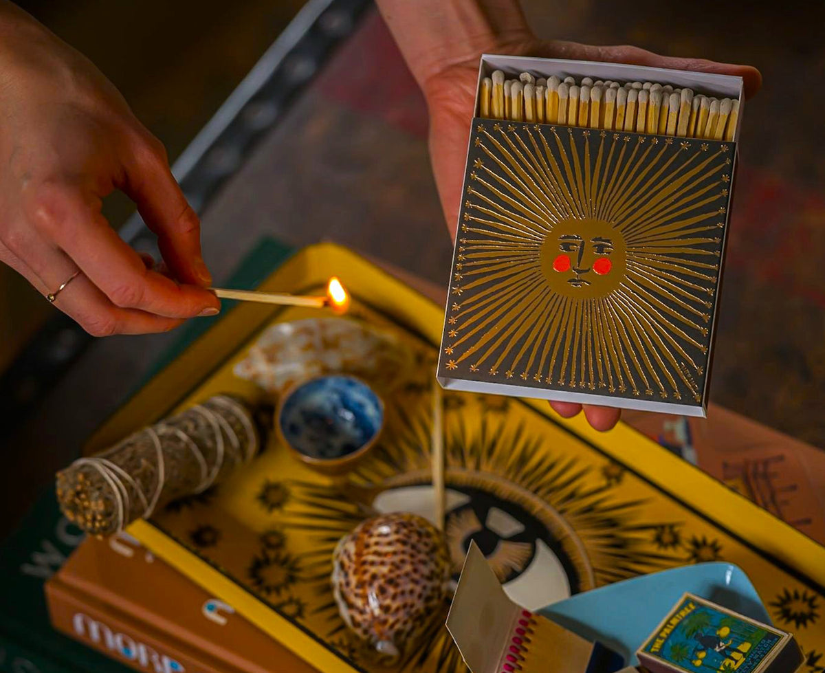 A matchbox with a black cover featuring a metallic gold sun design and red cheeks being used to light incense.