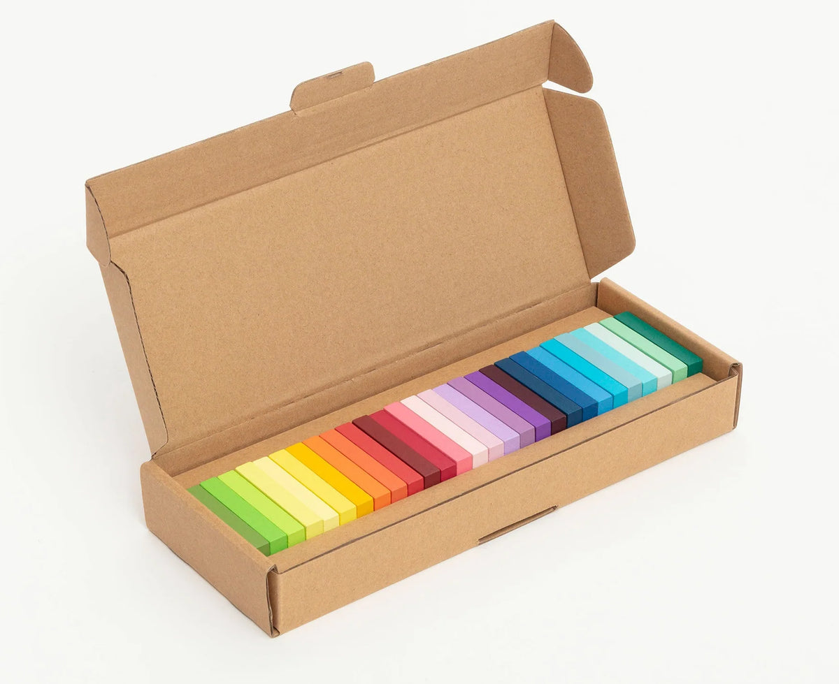 An open brown cardboard box containing a set of colorful rectangular dominoes arranged in a rainbow gradient, photographed on a clean white background.