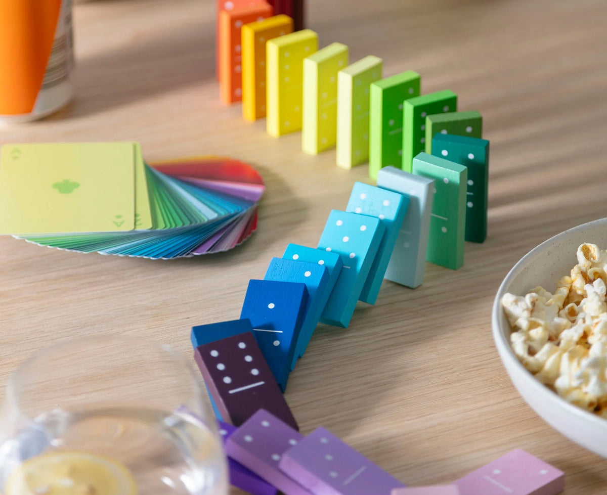 A colorful domino game arranged in a line on a light wood tabletop, with a fanned deck of cards and a bowl of popcorn nearby.