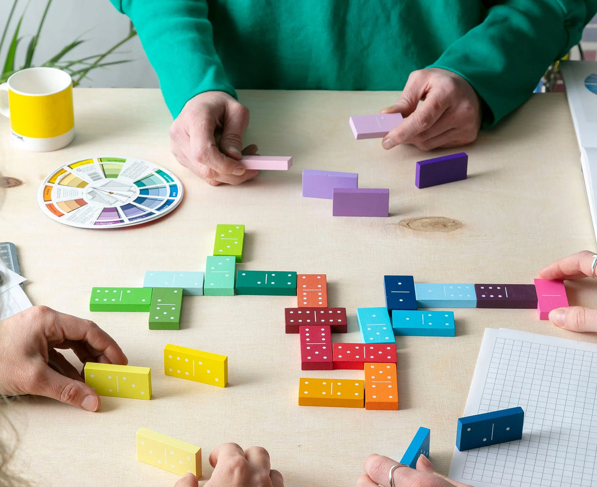 A group of people arranging colorful wooden domino tiles on a light wood table, with a color wheel, a yellow mug, and graph paper visible in a bright indoor setting.