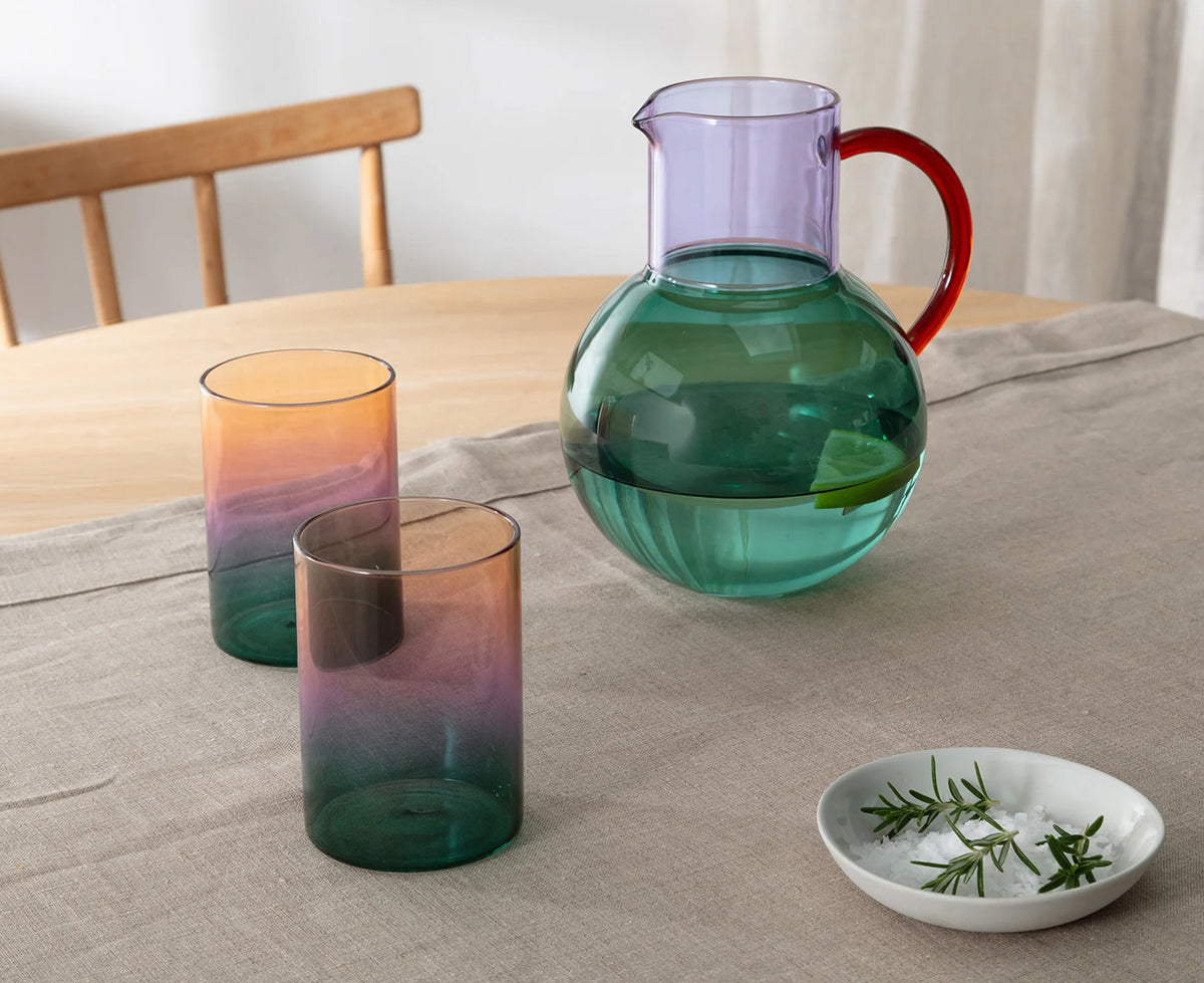 A set of three translucent glass tableware pieces on a linen tablecloth: a round green pitcher with a red handle and a purple neck, and two matching gradient tumblers with orange, purple, and green tones.