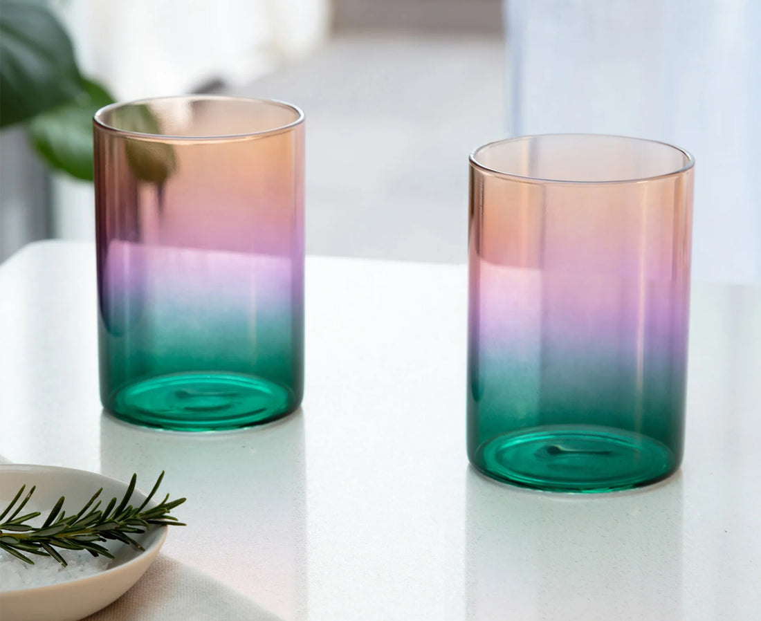 Two gradient glass tumblers with a color-shifting finish, transitioning from peach and pink to purple and deep teal, displayed on a white tabletop.