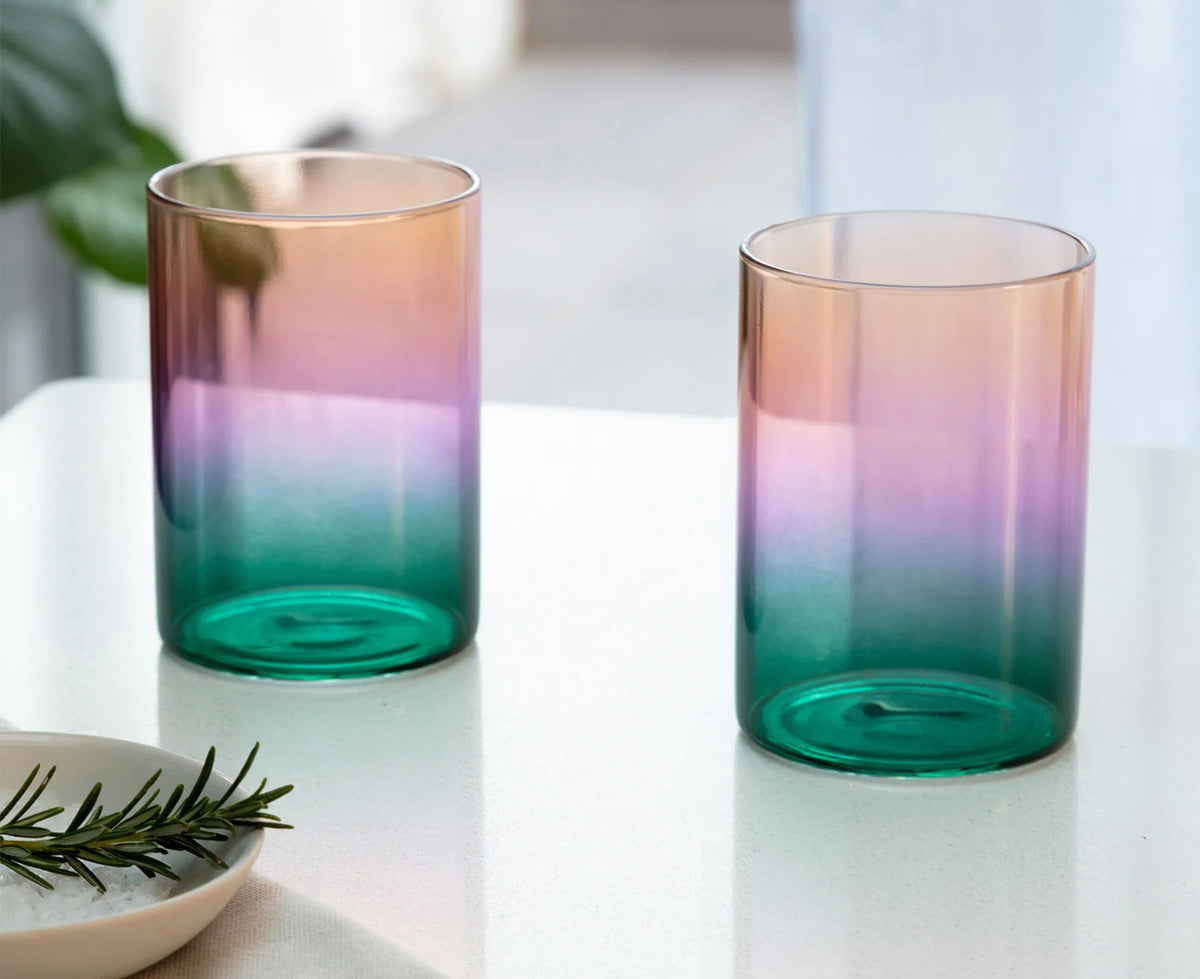 Two gradient glass tumblers with a color-shifting finish, transitioning from peach and pink to purple and deep teal, displayed on a white tabletop.