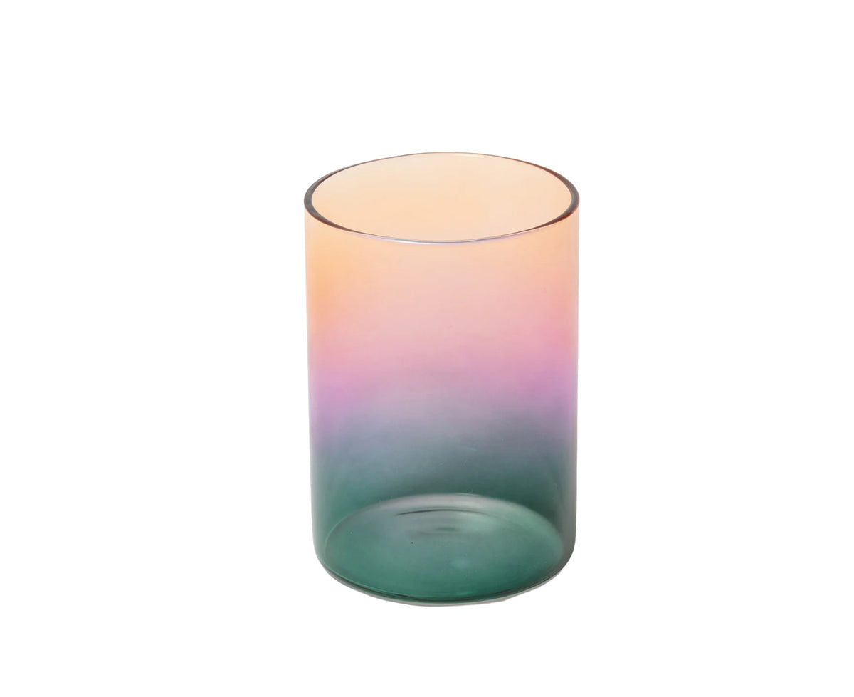 A cylindrical glass tumbler with a smooth ombre gradient transitioning from peach and pink at the top to purple and teal at the bottom, isolated on a white background.