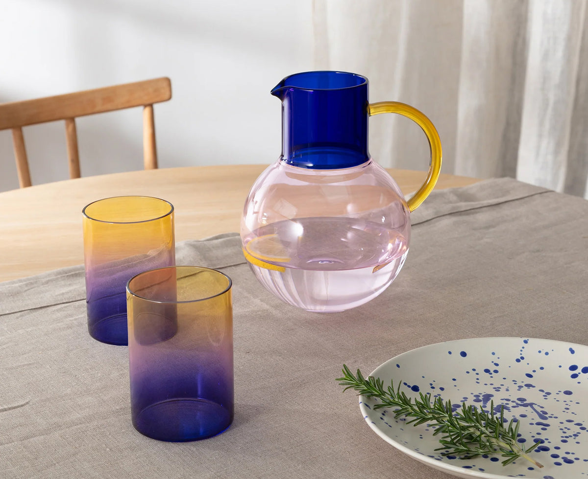 A set of glass tableware including a pink-tinted pitcher with a dark blue lid and yellow handle, two matching gradient tumblers, and a white speckled plate on a linen tablecloth.