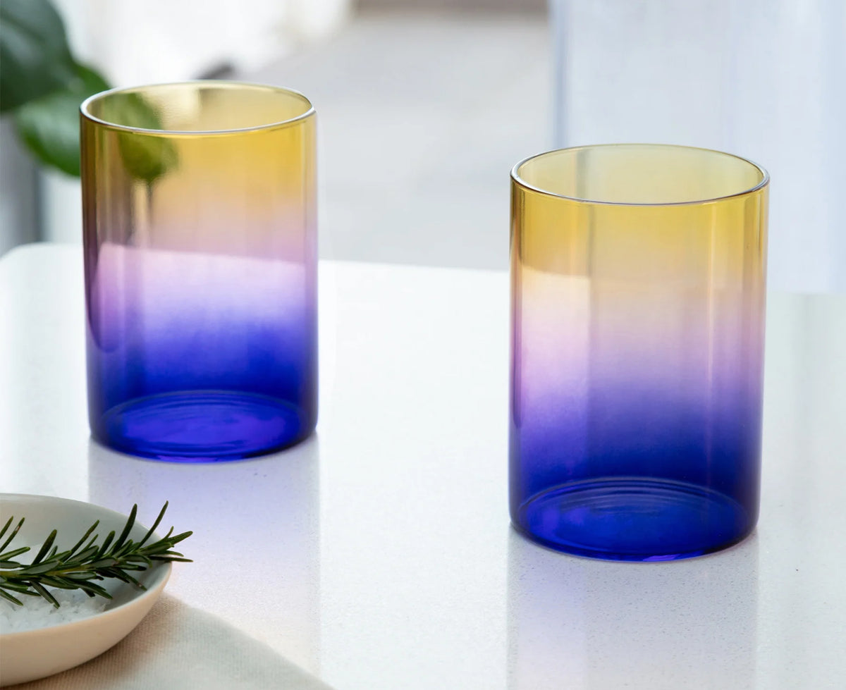Two gradient glass tumblers with a yellow-to-purple-to-blue color transition, displayed on a white tabletop with a rosemary sprig in a bowl.