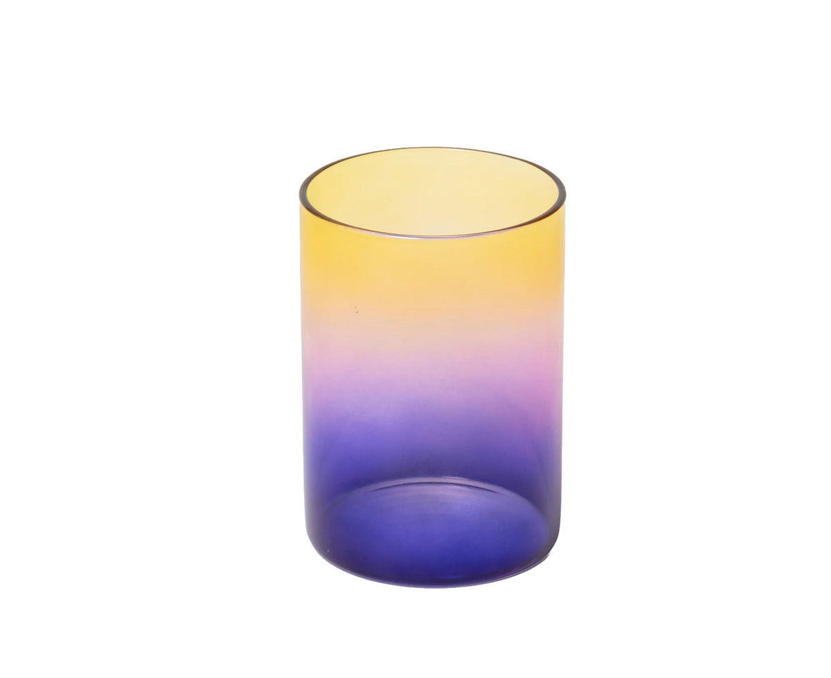 A translucent cylindrical glass tumbler with a smooth ombre gradient transitioning from yellow at the top to purple and deep blue at the bottom, isolated on a white background.