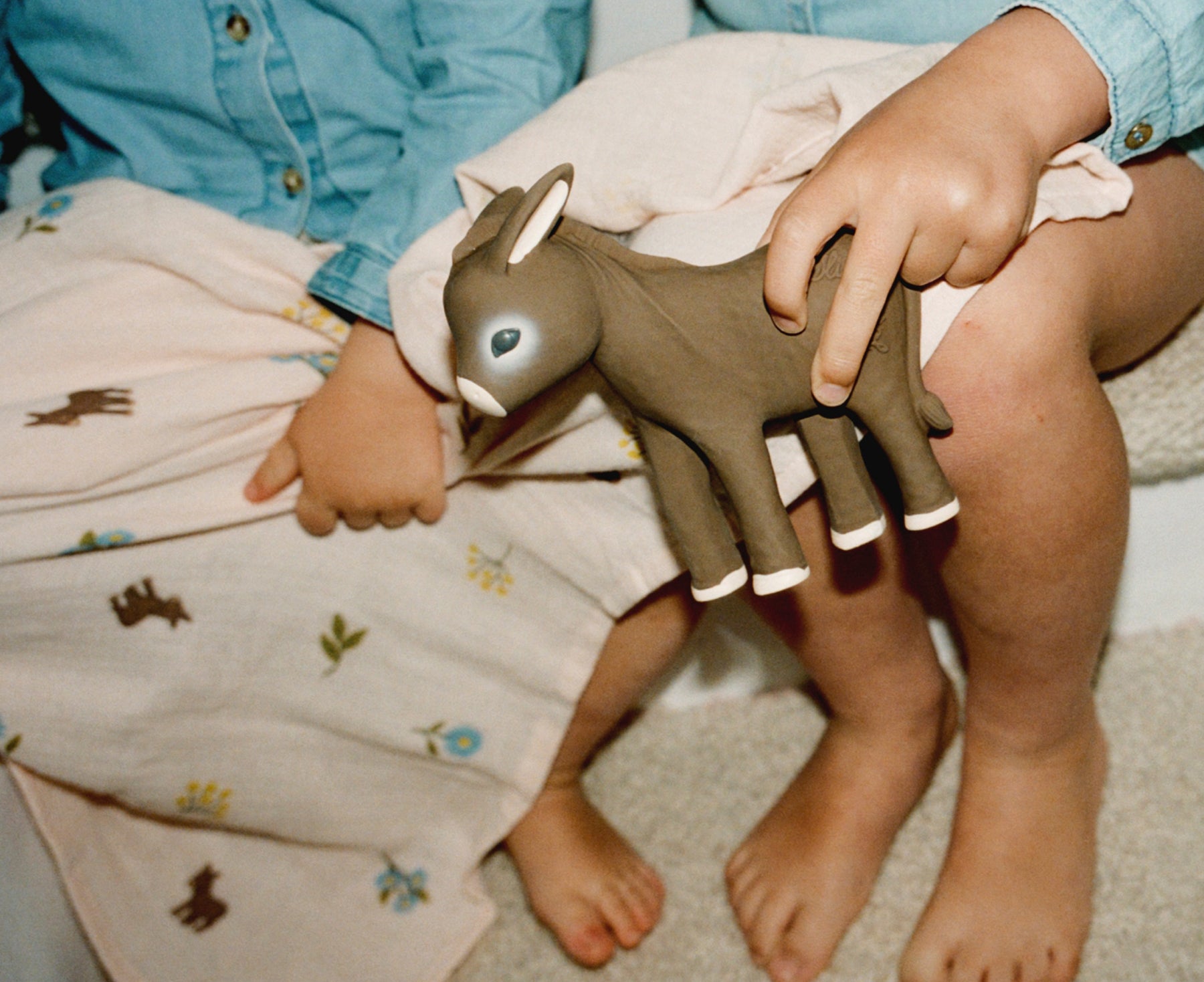 A child holding a Hector the Donkey Chewable Toy by Oli &amp; Carol sitting on a carpeted floor.
