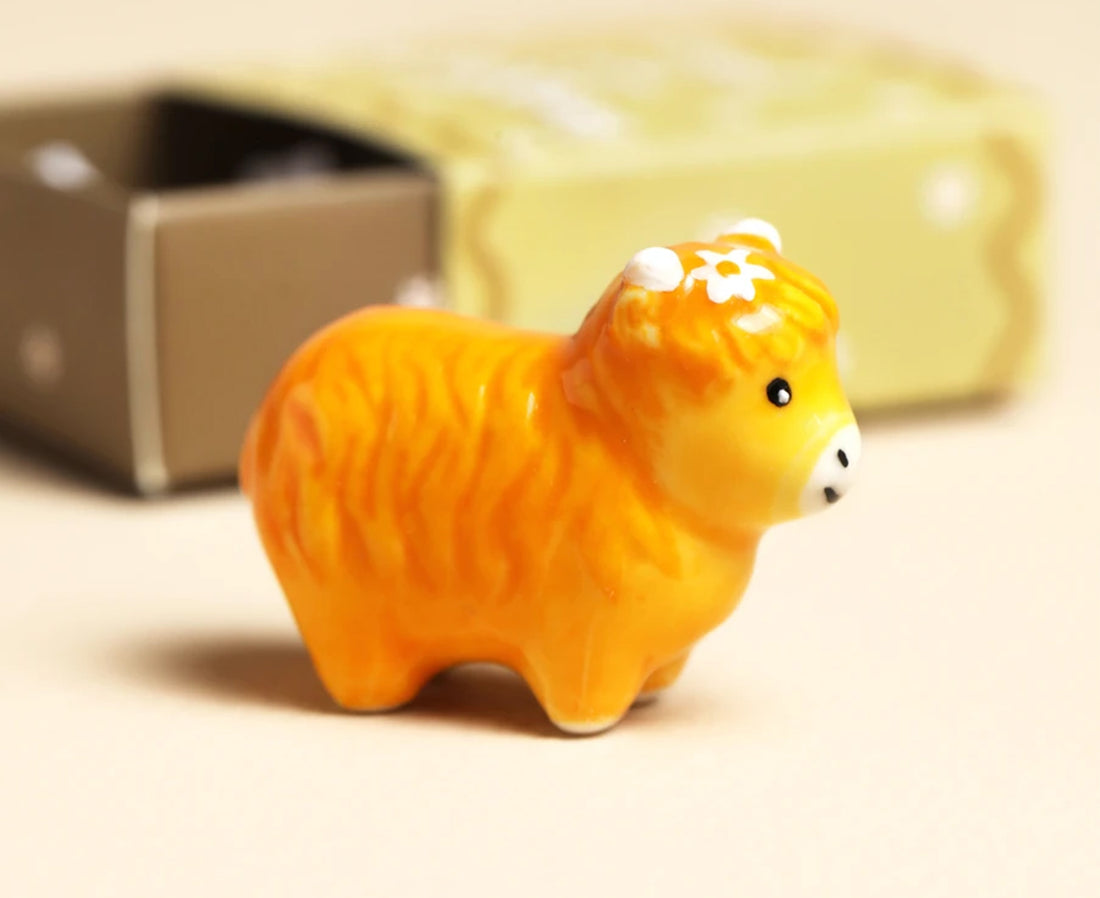 A small orange ceramic cow figurine with white accents and a flower on its forehead, standing in front of a blurred cream-colored box.