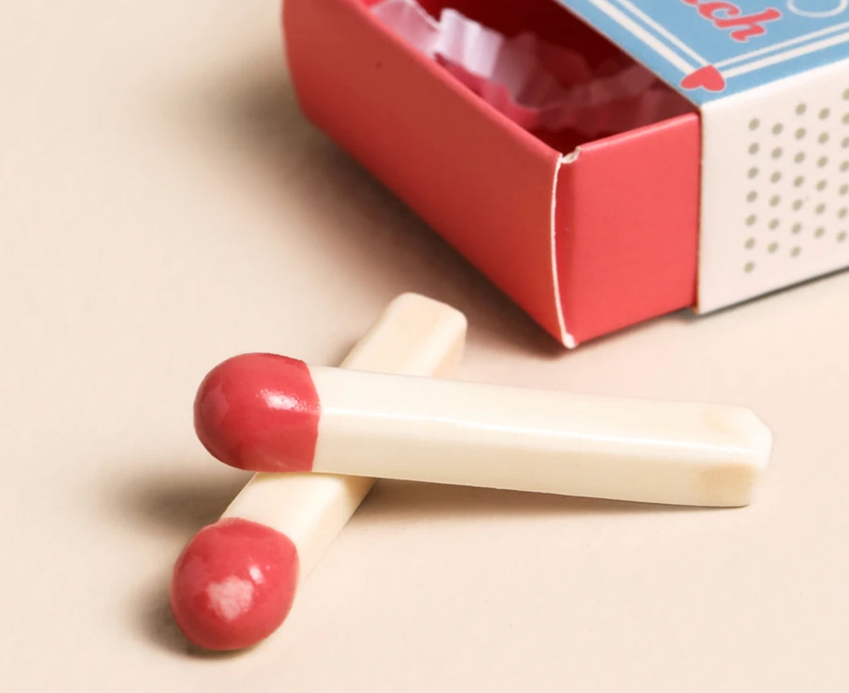 Two small ceramic matches with cream-colored handles and red tips are arranged in front of an open red and white matchbox on a light beige background.