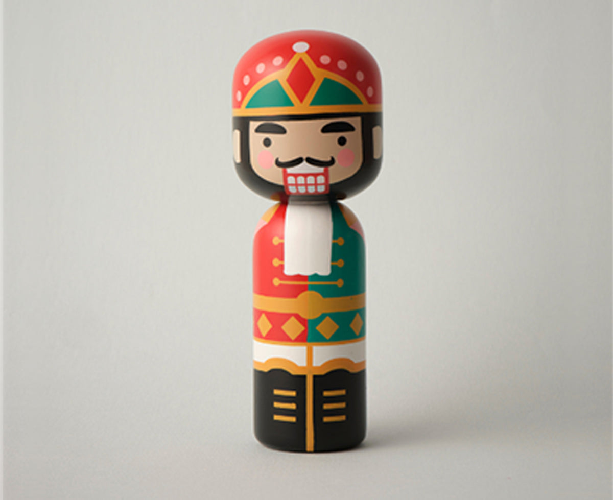 A colorful wooden nutcracker kokeshi doll with a red hat, green and red patterned coat, black pants, and a white beard, isolated on a gray background.