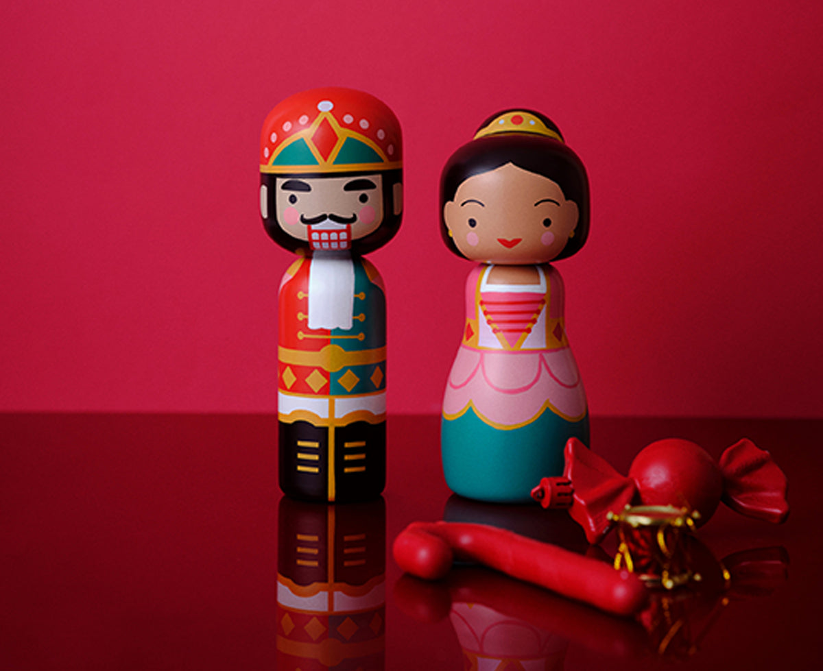 Two hand-painted wooden kokeshi dolls, a nutcracker-style figure in red and gold and a sugarplum-style figure in pink, red, and teal, standing next to a red lollipop and wrapped candy on a reflective red surface.