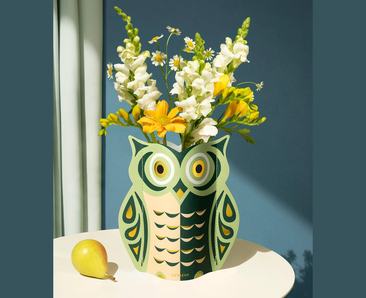 An owl-shaped paper vase featuring a stylized graphic design in green, peach, and gold by Octaevo. The vase holds yellow flowers and sits on a white table on a gray-blue background.