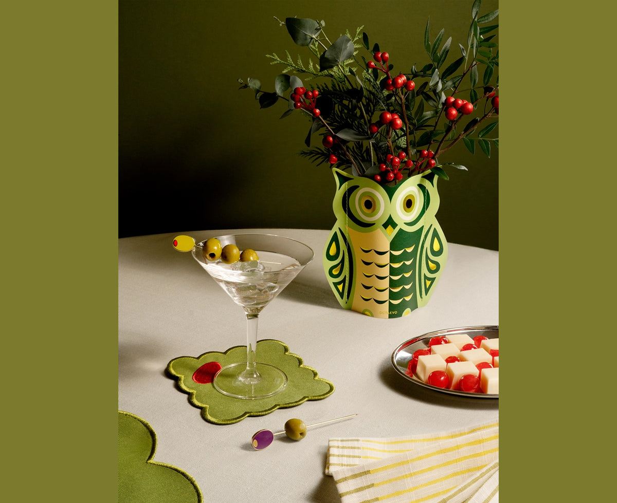 An owl-shaped paper vase featuring a stylized graphic design in green, peach, and gold by Octaevo. The vase holds holly with berries and sits next to a martini.
